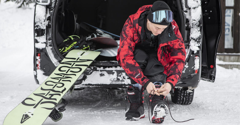 Mężczyzna przygotowuje się do zjazdu na snowboardzie siedząc przy otwartym bagażniku samochodu